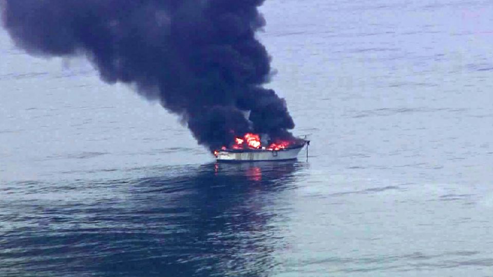 White boat engulfed in black smoke and flames on the water