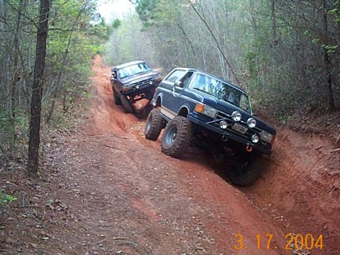 1986 Ford Bronco - Two off-road vehicles navigate a steep, muddy trail through a wooded area.