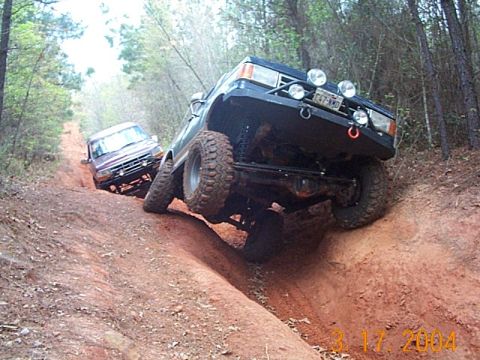 1986 Ford Bronco - Two SUVs navigate a steep, muddy incline on a wooded trail, their off-road capabilities.