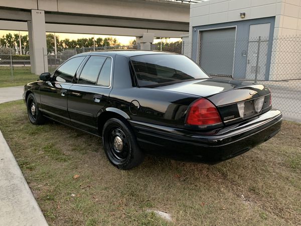 Black Ford Crown Victoria sedan with black steel wheels, rear three-quarter view, parked near a fence and overpass