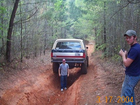 1986 Ford Bronco - A lifted Ford Bronco navigates a narrow dirt path in a wooded area, observed by two onlookers.