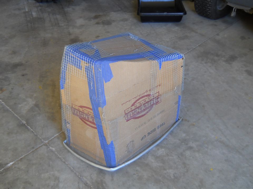 Cardboard box covered in wire mesh and blue tape, with a metal base, on a concrete floor
