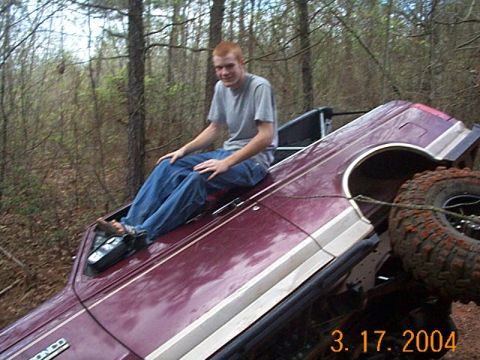 1986 Ford Bronco - A man sits atop a maroon SUV that has rolled over in a wooded area.