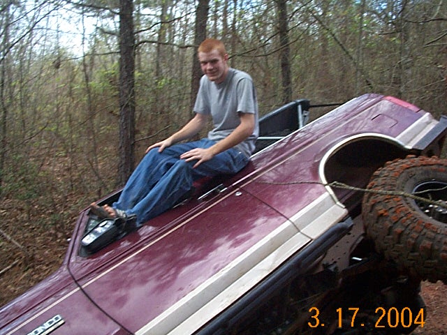 Maroon 1980s SUV overturned in woods, with a man sitting on its roof