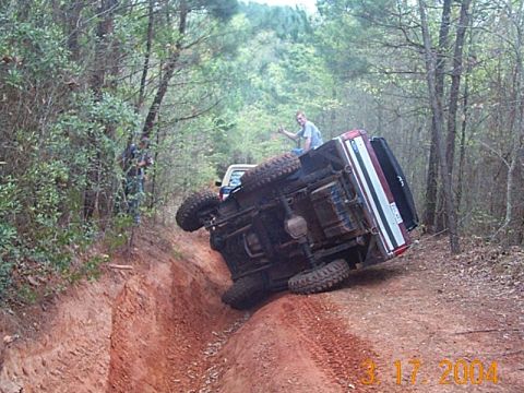1986 Ford Bronco - A 1986 Ford Bronco has rolled onto its side while navigating an off-road trail, with another vehicle visible in the background.