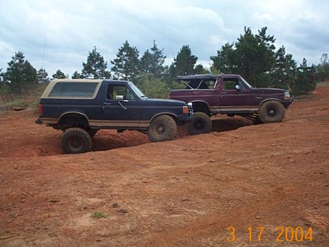 1986 Ford Bronco - Two Ford Broncos are shown navigating a dirt path in a wooded area under a cloudy sky.