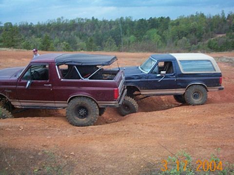 1986 Ford Bronco - Two Ford Broncos navigate a dirt trail, one with a removable top and the other with a hard shell.