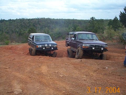 1986 Ford Bronco - Two modified Ford trucks, a purple F-150 and a blue Bronco, tackle an off-road trail with significant suspension articulation.
