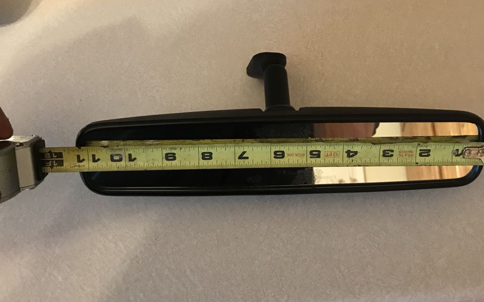 Measuring a black rear-view mirror with a tape measure. The tape shows about 10.5 inches