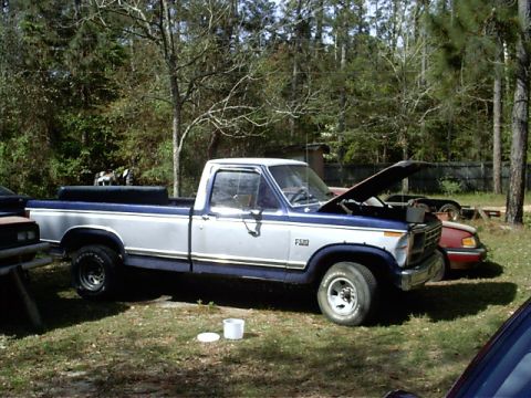 1985 Ford Bronco - A two-tone Ford F-150 pickup truck is parked outdoors with its hood up, next to another vehicle.