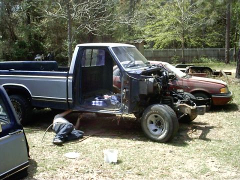 1985 Ford Bronco - A person works on the underside of a pickup truck with its hood and doors open, while another vehicle is parked behind it.