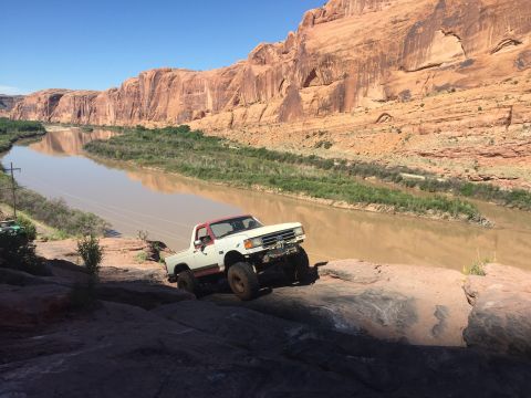 1984 Ford Bronco - A modified pickup truck navigates a rocky trail beside a wide, muddy river under a clear blue sky.