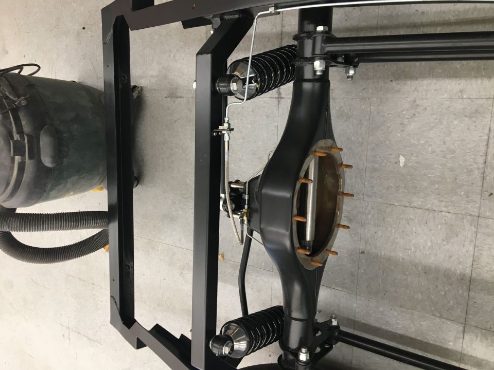 Close-up of a rear axle assembly with shocks and springs, mounted on a black frame. A large green tank is to the left