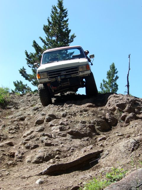 1984 Ford Bronco - A white Ford Bronco navigates a steep, rocky trail under a clear blue sky.
