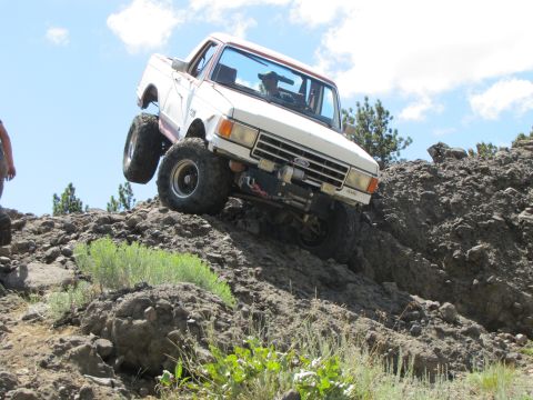 1984 Ford Bronco - This lifted pickup truck navigates a steep, rocky incline under a partly cloudy sky.