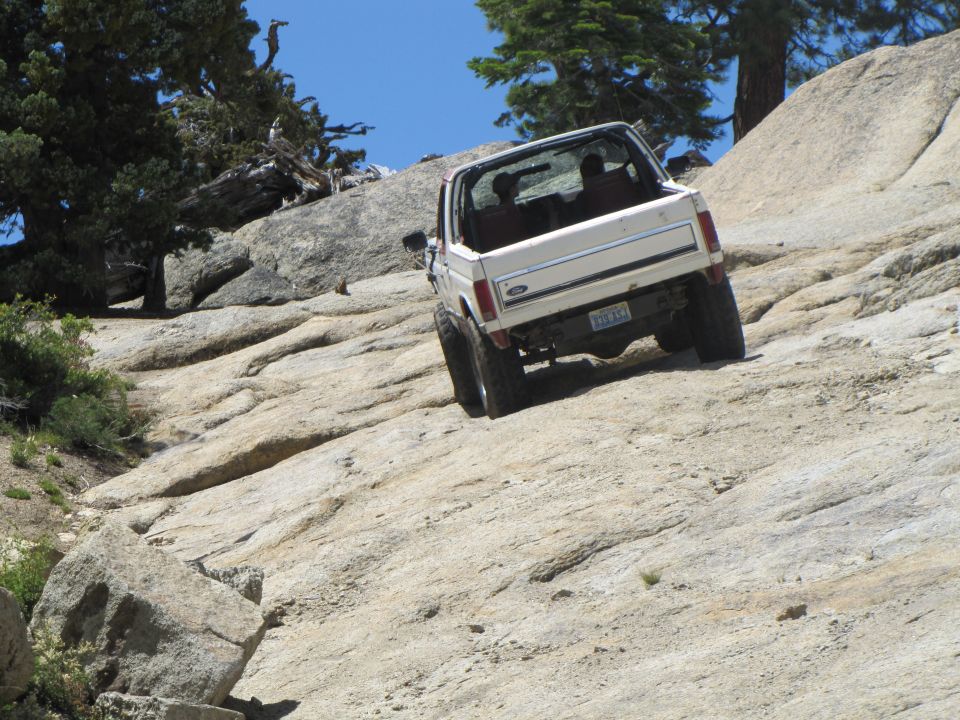 White 1984 Ford Bronco climbing a steep, rocky incline with trees and blue sky in the background
