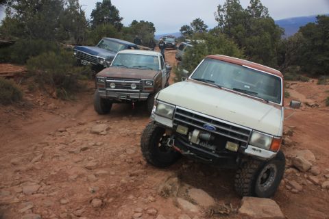 1984 Ford Bronco - Several off-road vehicles navigate a rough, rocky path through a wooded, arid landscape.