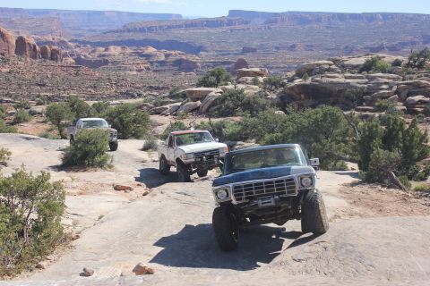 1984 Ford Bronco - Three modified Ford trucks navigate a rocky, off-road trail in a desert landscape.