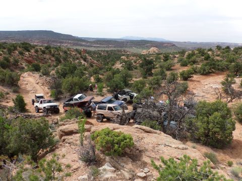1984 Ford Bronco - Several off-road vehicles are gathered on a rocky trail surrounded by desert vegetation and hills.