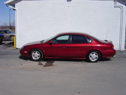 1998 Ford Taurus - Side view of a red 1998 Ford Taurus sedan parked in front of a white building.
