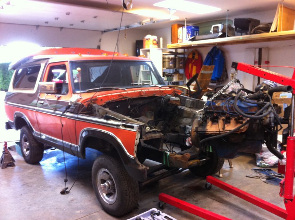 Orange and brown 1978 Ford Bronco in a garage, engine removed and suspended by a hoist