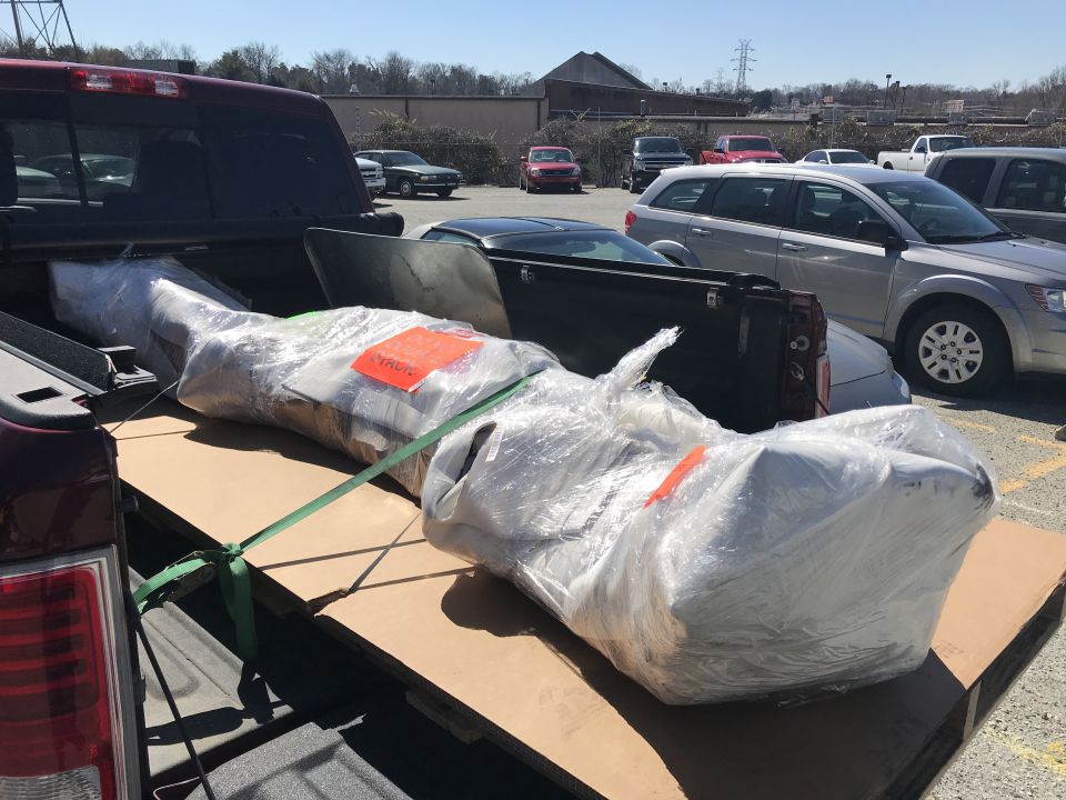 Wrapped car part secured with straps in the bed of a pickup truck, with other vehicles parked in the background