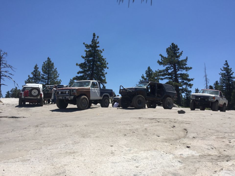Several modified off-road trucks and SUVs parked on a dusty, rocky surface under a clear blue sky