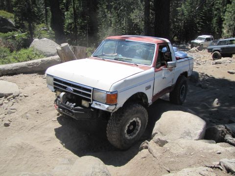 1984 Ford Bronco - A modified Ford F-Series truck drives off-road with other vehicles in the background.