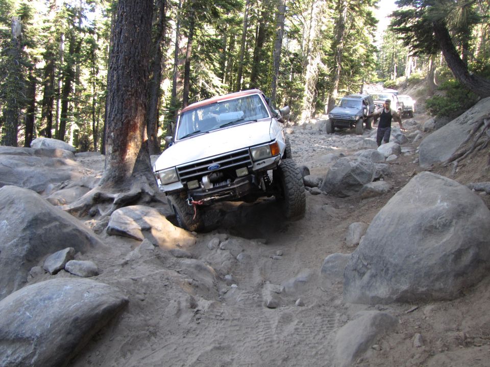White off-road SUV with a red top navigating a rocky trail, other vehicles behind
