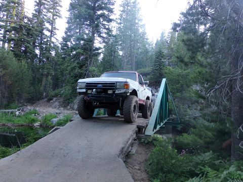 1984 Ford Bronco - A white pickup truck with large off-road tires navigates a narrow bridge in a wooded area. The vehicle's body style is indicative of a 1980…