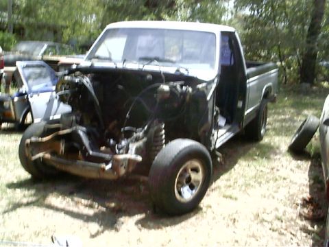 1985 Ford Bronco - This pickup truck is missing its hood and engine, revealing its front suspension and chassis. A spare tire lies nearby.