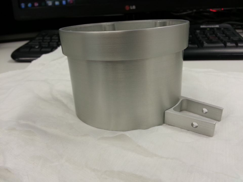 Silver metal cylindrical part with bracket on white cloth, blurred keyboard and monitor in background.
