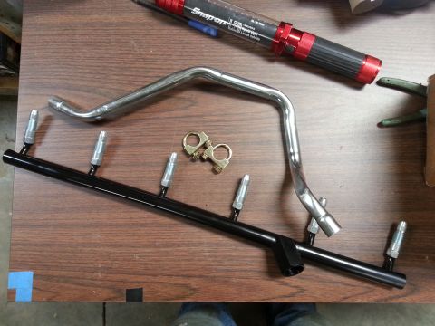 1978 Ford Bronco - A black fuel rail with four injectors is laid out on a wooden table next to a bent chrome pipe and a Snap-On flashlight.