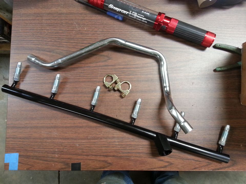 Black fuel rail with injectors and chrome pipe on wooden table.