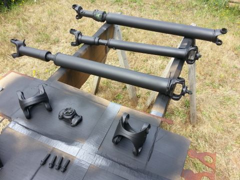 1978 Ford Bronco - Three black driveshafts are laid out, with two U-joints visible on each. Several black metal parts, including a yoke and some bolts, are on…