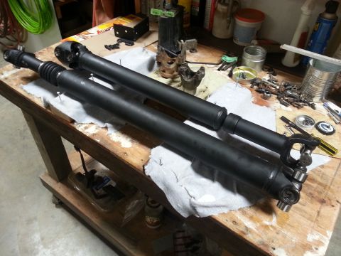 1978 Ford Bronco - Two black driveshafts are laid out on a wooden workbench, resting on white cloths. Various tools and hardware are scattered around the driv…
