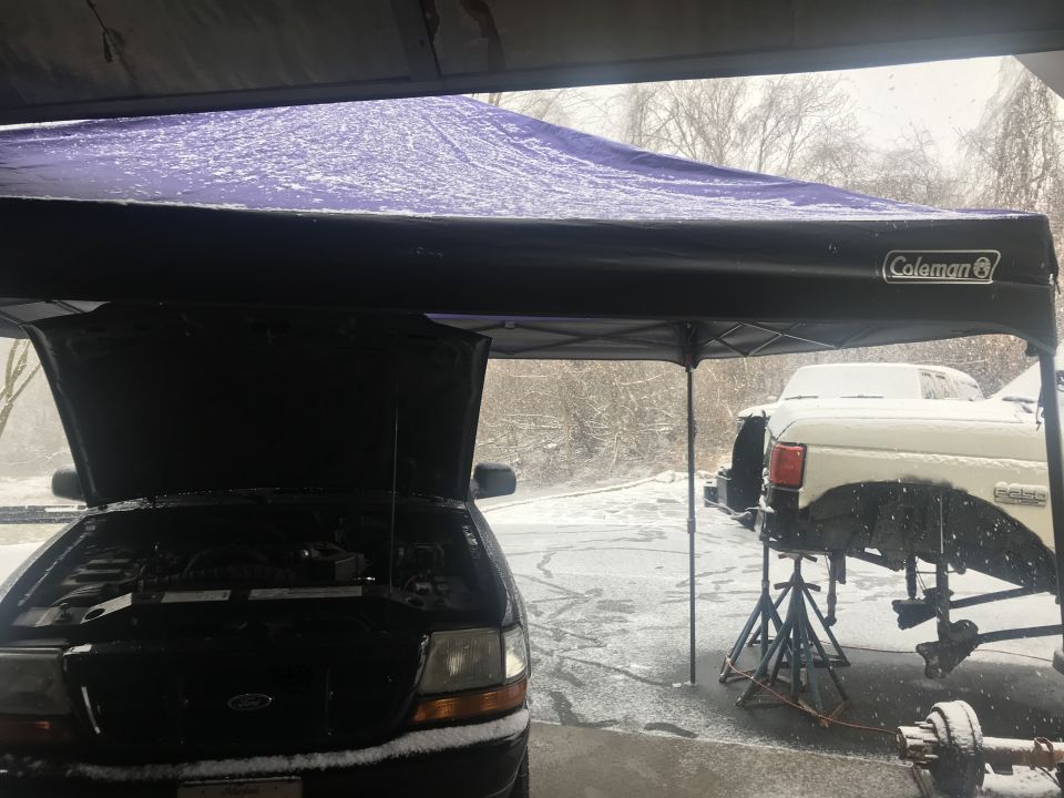 Black Ford Ranger with hood open under a purple canopy during snowfall. White Ford truck on jack stands.