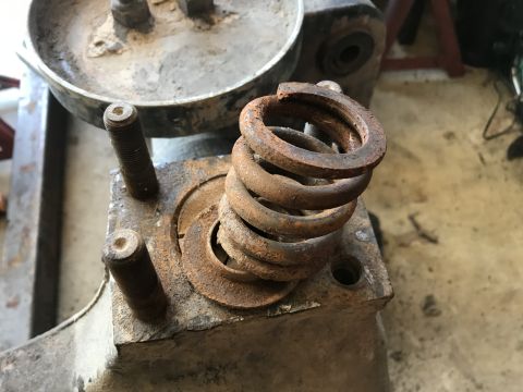1989 Ford Bronco - A rusted coil spring sits atop a metal component with two threaded bolts protruding from its surface. The background shows other vehicle pa…