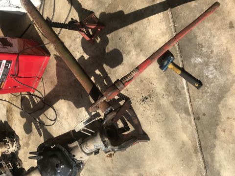 1989 Ford Bronco - A red and yellow jack is positioned over a rusty differential housing. A red welder and a hammer are also visible on the concrete floor.