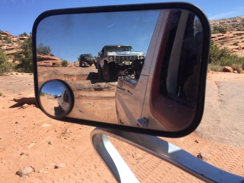 1984 Ford Bronco - A reflection in a side mirror shows two off-road vehicles navigating a rocky, desert terrain. The main mirror also reflects the landscape a…
