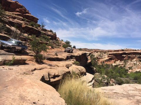1984 Ford Bronco - A blue and silver 1980s Ford Bronco drives on a rocky, off-road trail in a desert landscape with red rock formations and a bright blue sky.