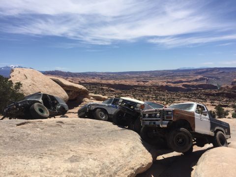 1984 Ford Bronco - Three modified off-road vehicles are parked on rocky terrain with a vast desert landscape and mountains in the background under a blue sky…
