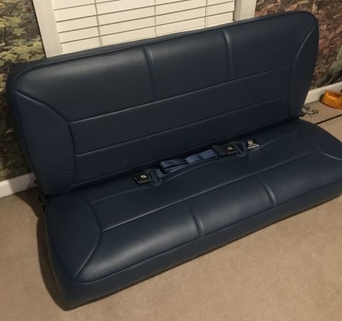 1995 Ford Bronco - A dark blue bench seat with integrated seat belts is shown from a slightly elevated angle, resting on a carpeted floor. The seat appears to…