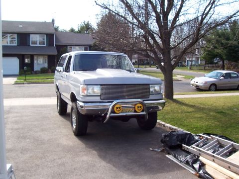 1990 Ford Bronco - A silver Ford Bronco is parked in a suburban driveway, showing its front grille and hood scoop.