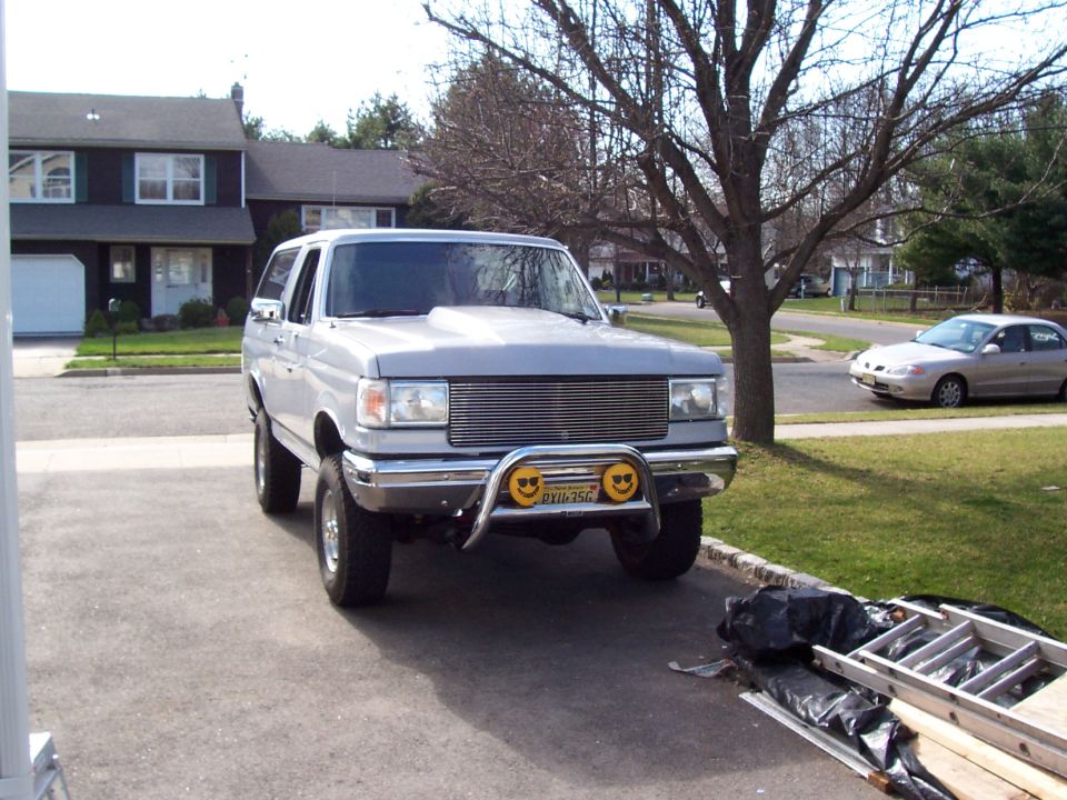 Silver Ford Bronco parked in driveway, front view. Has a chrome grille guard with smiley face lights and a hood scoop
