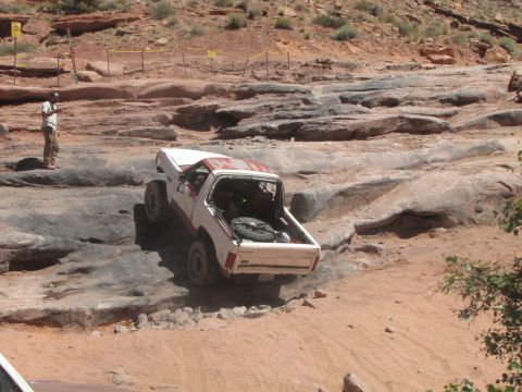1984 Ford Bronco - A white and red Ford Bronco is navigating a rocky, off-road trail with a person standing nearby observing. The vehicle is positioned on a d…