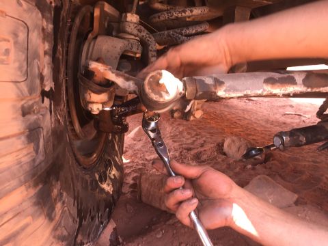 1984 Ford Bronco - A person is using a wrench to tighten a tie rod end on the front suspension of a vehicle, likely an off-road vehicle given the sandy terrai…