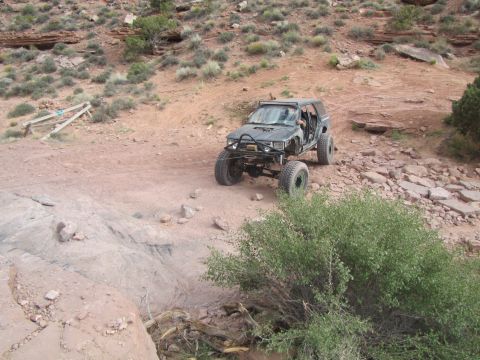 1984 Ford Bronco - A modified dark-colored SUV is navigating a rocky, off-road trail with a winch line attached. The vehicle appears to be in a desert environ…