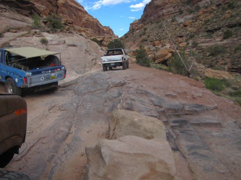 1984 Ford Bronco - Two off-road vehicles are driving on a rocky trail through a canyon. The lead vehicle is a blue pickup truck with a canvas cover over the b…