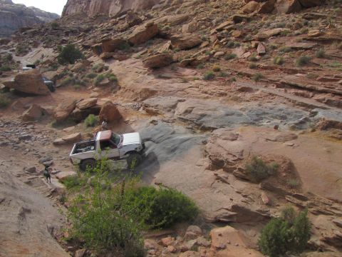 1984 Ford Bronco - A white and red Ford pickup truck navigates a rocky, off-road trail in a desert canyon. The truck is angled on a steep, rocky incline with…